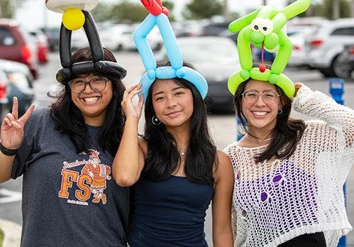 Three smiling young women wearing balloon animal hats, standing in a parking lot.