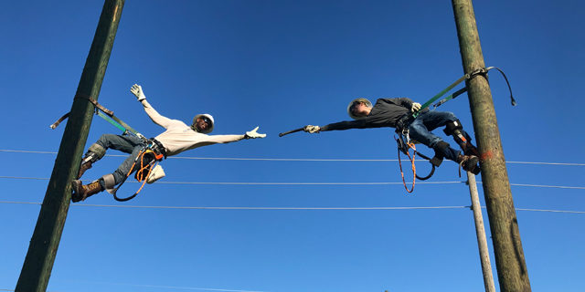 Two men climbing utility poles and handing something across to one another with a deep blue sky background