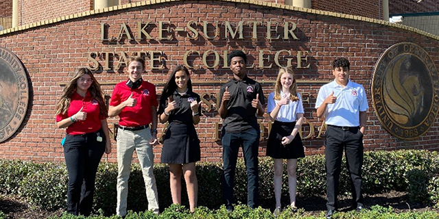 Group of students standing in front of brick Lake-Sumter State College sign showing thumbs up